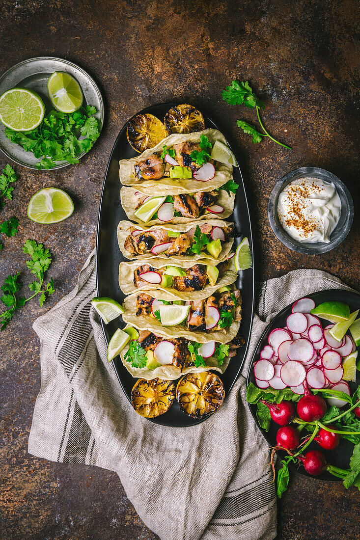 Gegrillte Hähnchen-Tacos auf einer dunklen ovalen Platte mit geschnittenen Radieschen, Limetten, Koriander, verkohlten Zitronen und saurer Sahne