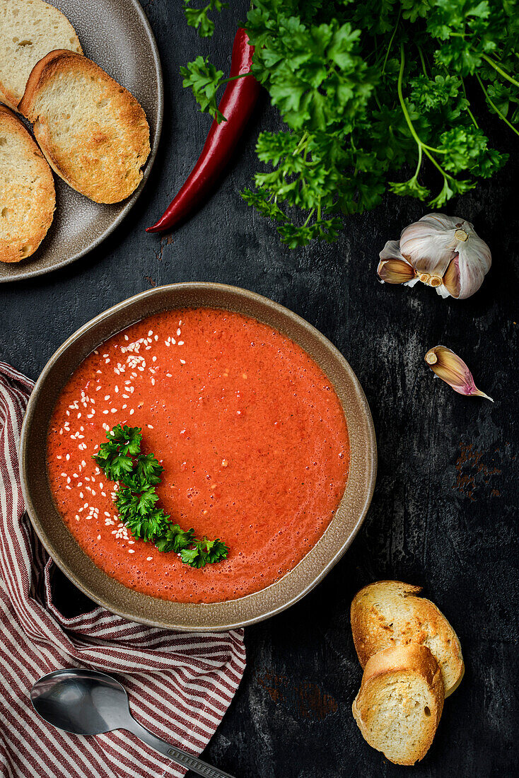 Gazpacho garnished with parsley in a wavy-edged plate. On the table are red peppers, tomatoes, garlic, croutons and a spoon. View from above