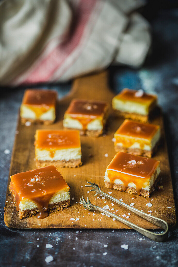 Small cheesecake squares with caramel sauce and sea salt on a wooden board, with small tongs