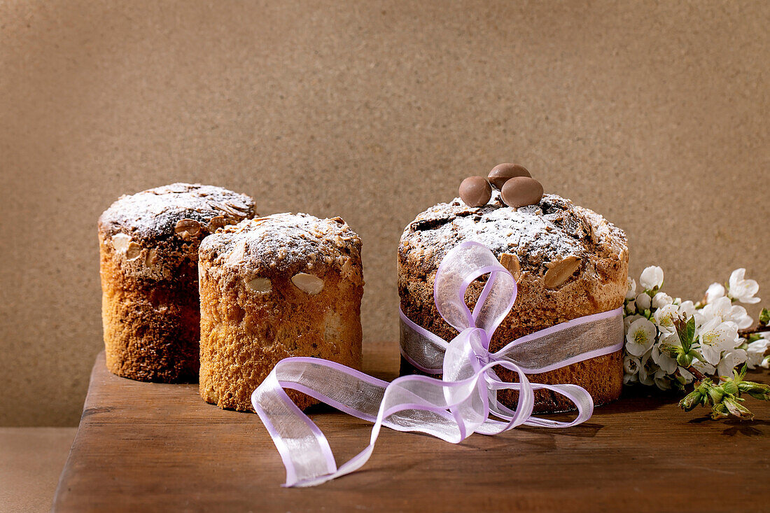 Homemade traditional Italian Easter panettone cake in a row, decorated with chocolate eggs, pink ribbon and blooming cherry blossoms on a wooden table. Traditional European pastry. Place to copy