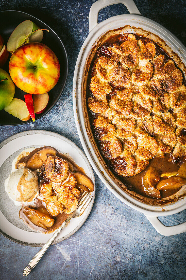 Apple cobbler in a ceramic baking dish, served on a plate with vanilla ice cream and a bowl of fresh apples