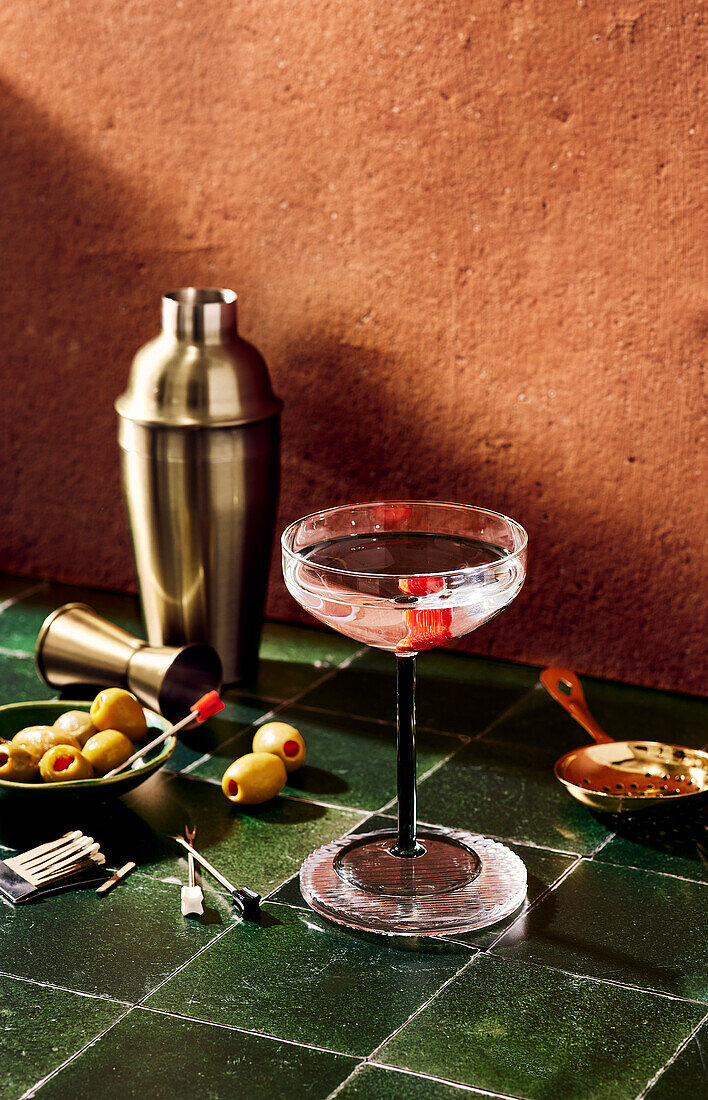 A cocktail glass and cocktail shaker on a bar with olives
