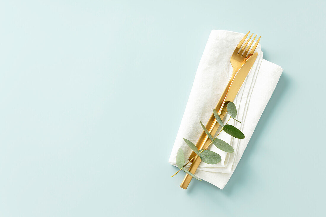 Golden cutlery with eucalyptus branches on a white linen napkin against a blue background. Minimalist design. Space for copying