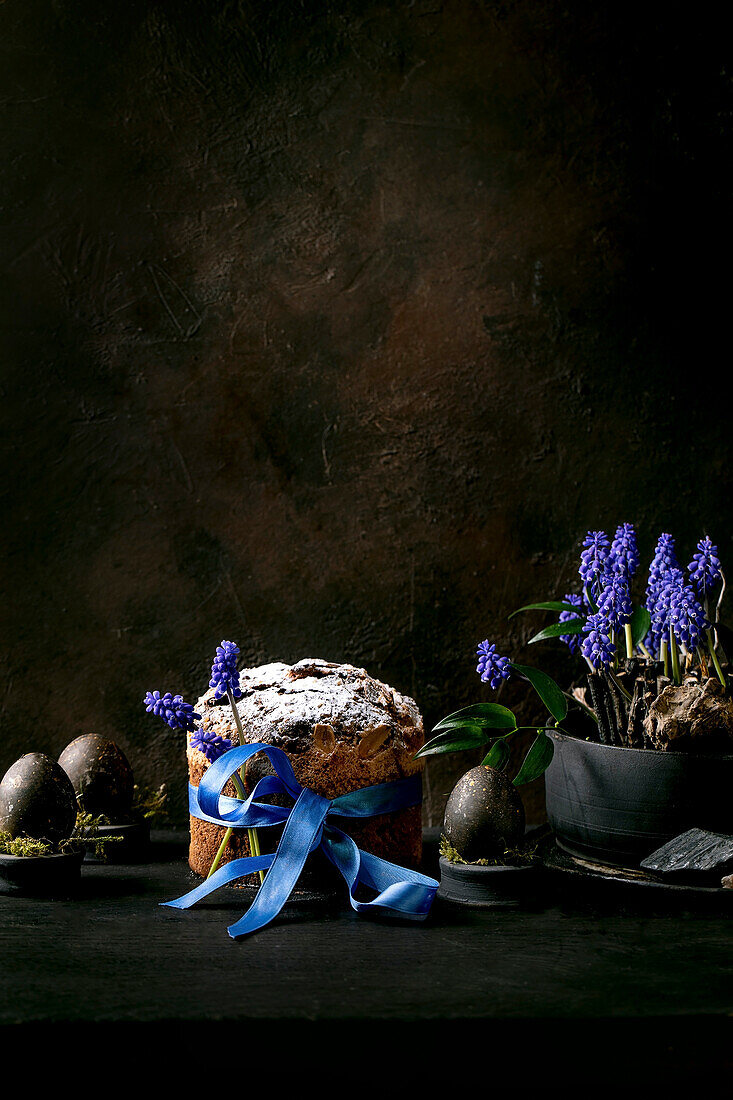 Hausgemachter traditioneller Oster-Panettone-Kuchen mit blauem Band, gefärbte schwarze Eier in Moos, blühende Muscari-Blumen auf schwarzem Holztisch. Traditionelle italienische Ostern backen, kopieren Raum
