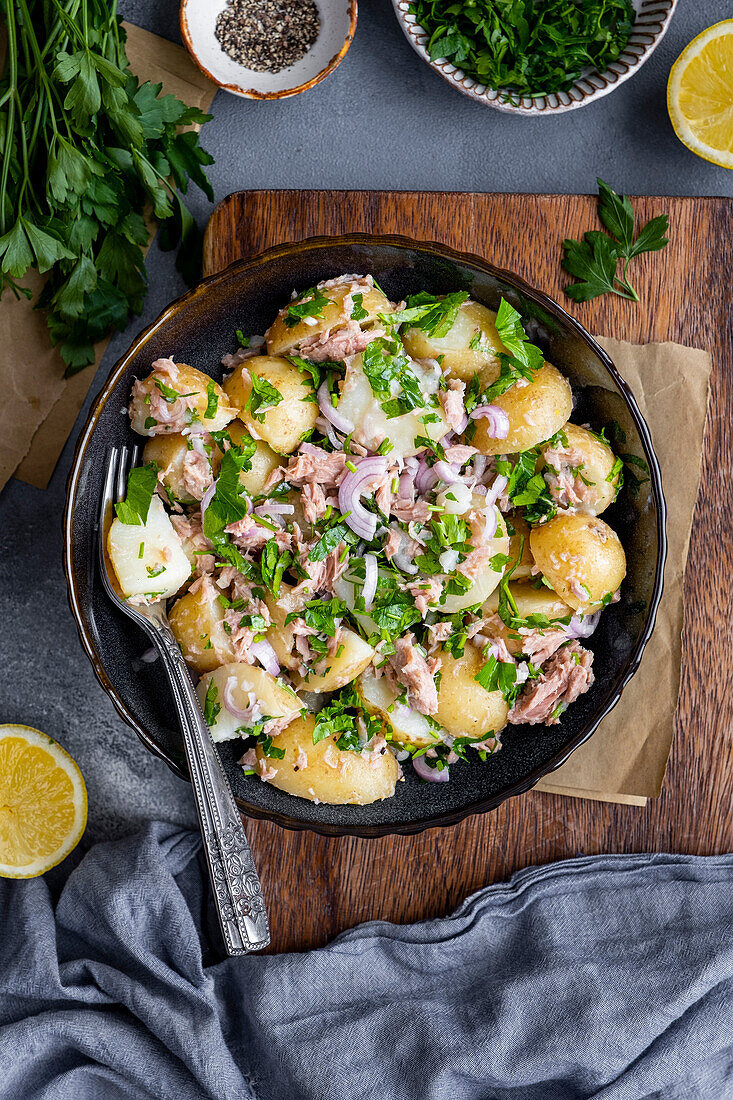 Tuna and potato salad in a black bowl with a fork, lemon halves, parsley and black pepper on the side