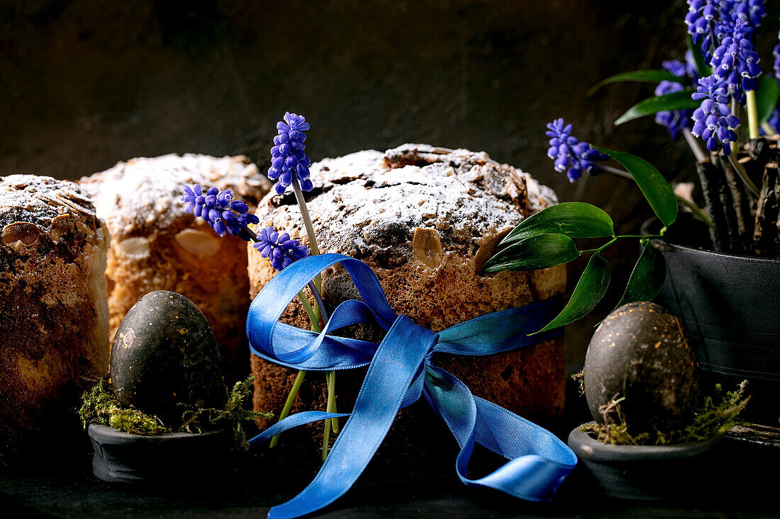 Hausgemachter traditioneller Oster-Panettone-Kuchen mit blauem Band, gefärbten schwarzen Eiern, blühenden Muscari-Blumen auf schwarzem Holztisch. Traditionelle italienische Osterbäckerei, Kopierraum, Nahaufnahme