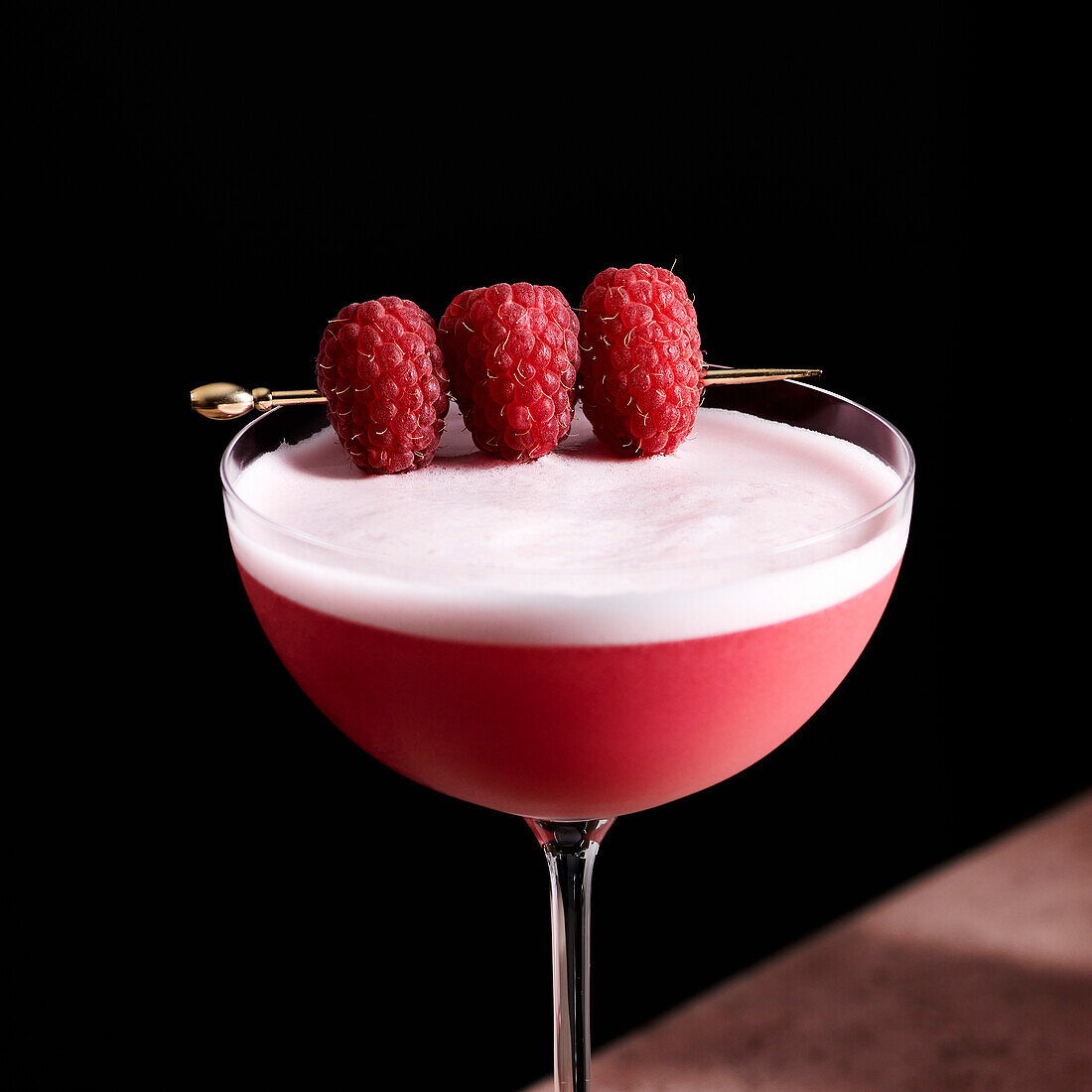 Clover club cocktail in a tall coupe glass garnished with three raspberries on a gold pick.