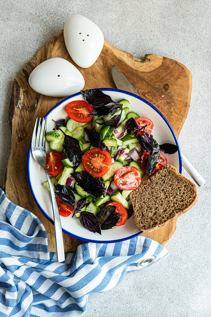 Draufsicht auf einen saisonalen Gemüsesalat mit Bio-Gurke, Tomate, Zwiebel und roten Basilikumblättern, serviert auf einem Teller mit Besteck auf einem Holztablett neben einer Serviette vor grauem Hintergrund