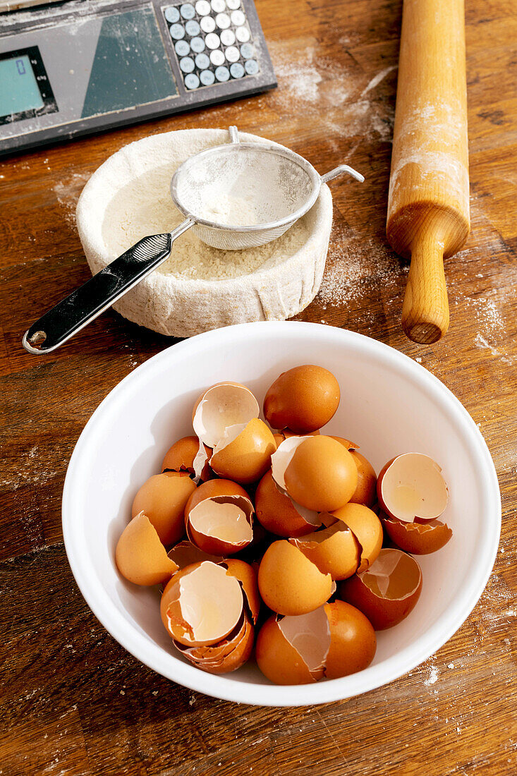 High angle view of flour with eggshells in bowls and sieve with rolling pin placed on wooden table for making pastry dishes in bakehouse