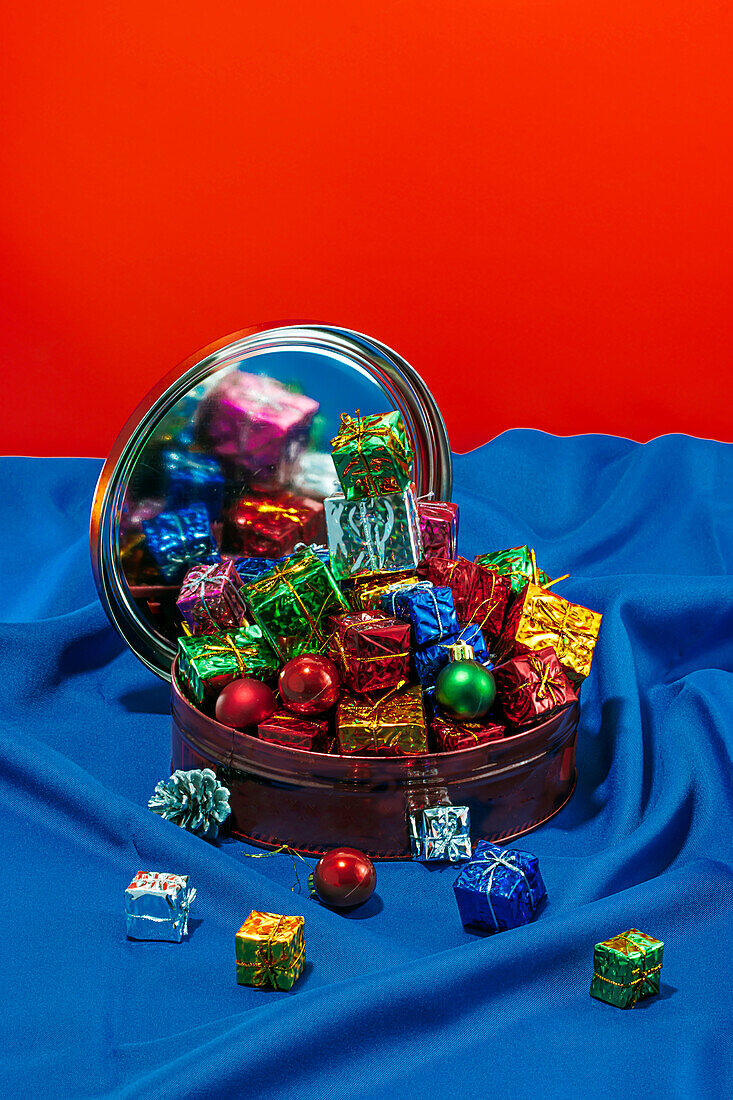 A tin filled with shiny Christmas candies and baubles reflected in a round mirror on a blue cloth