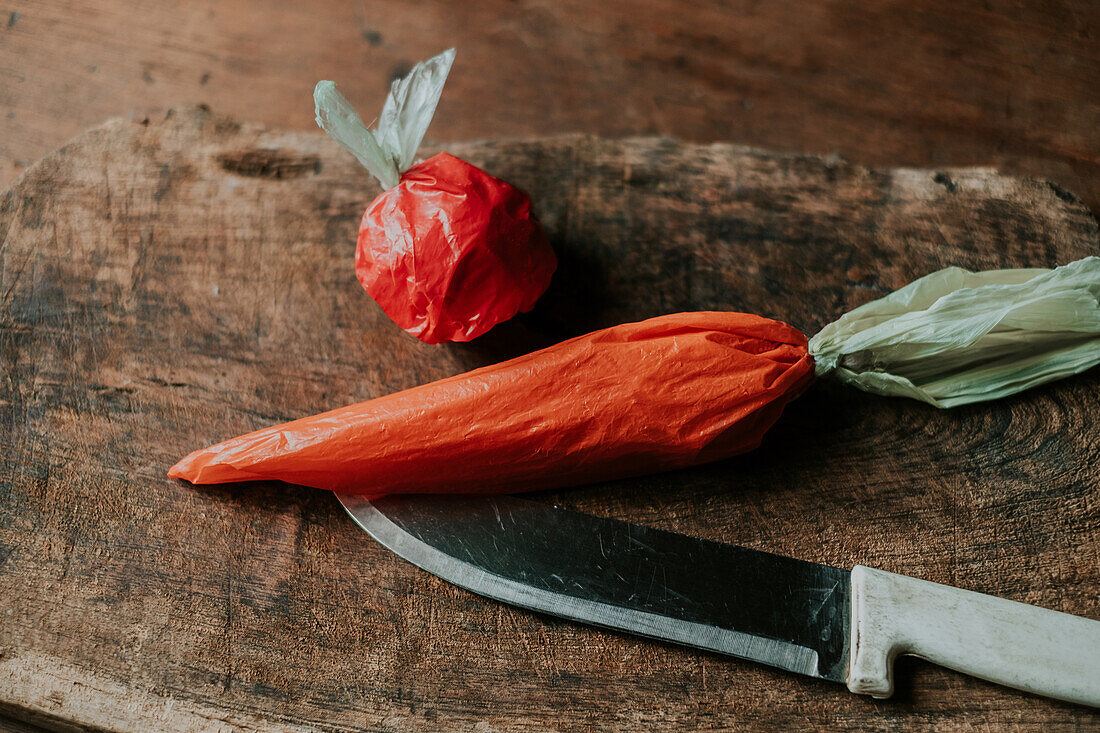 A red plastic bag shaped like a tomato and an orange bag resembling a carrot next to a knife on a wooden board