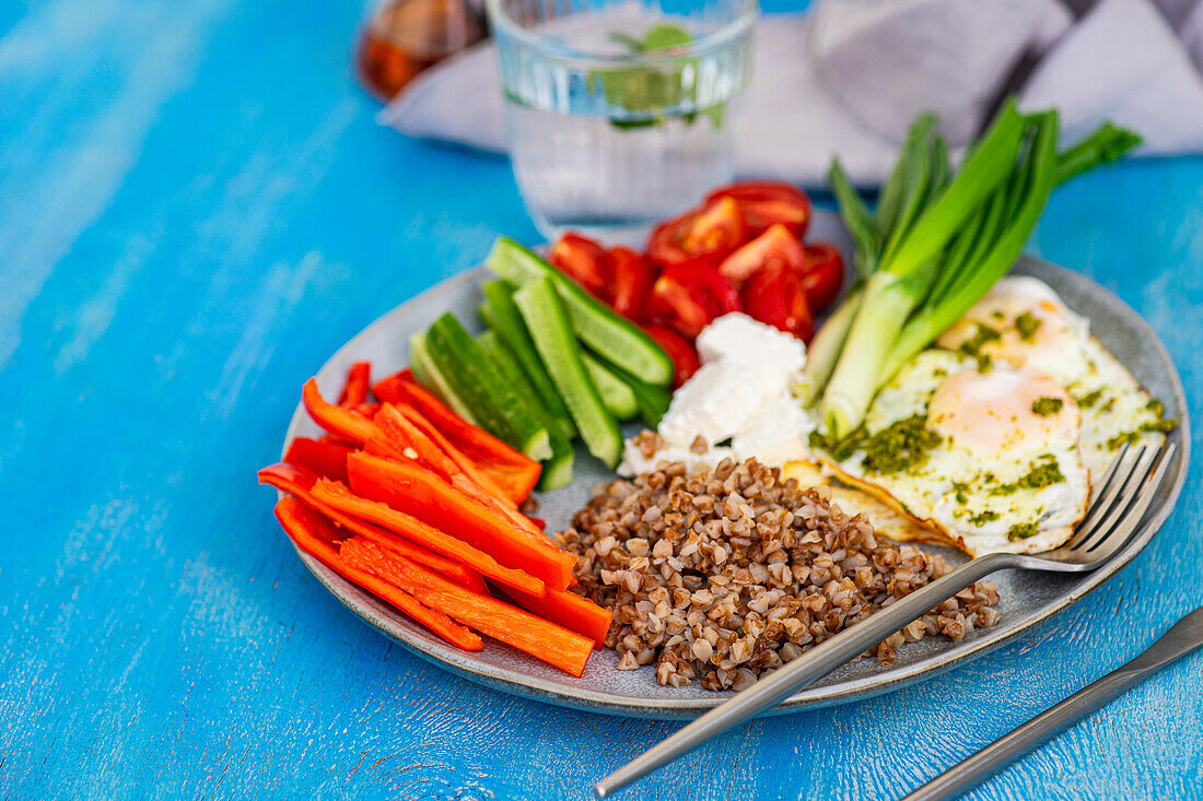 A nutritious breakfast setup showcasing sliced raw vegetables, feta cheese, buckwheat, and fried eggs with pesto on a blue background.