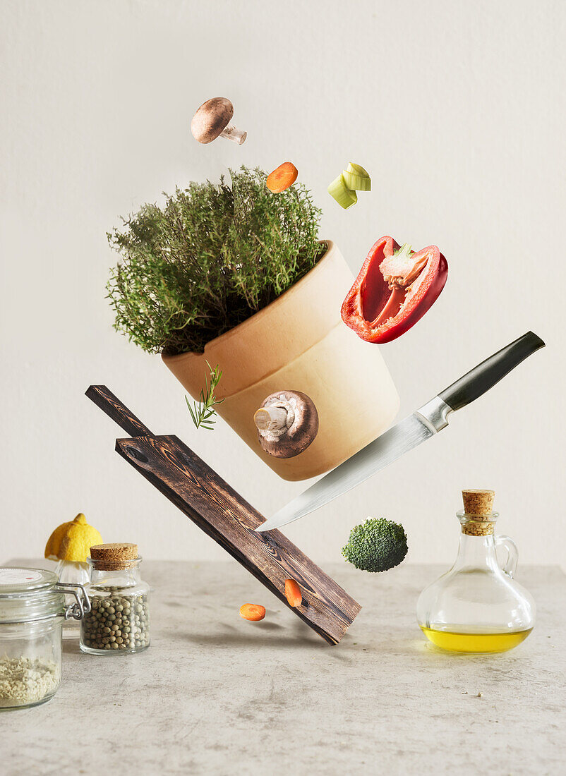Creative food levitation concept with flying cutting board, knife, vegetables, potted herbs and utensils at grey concrete kitchen table. Cooking at home with flavorful and healthy ingredients