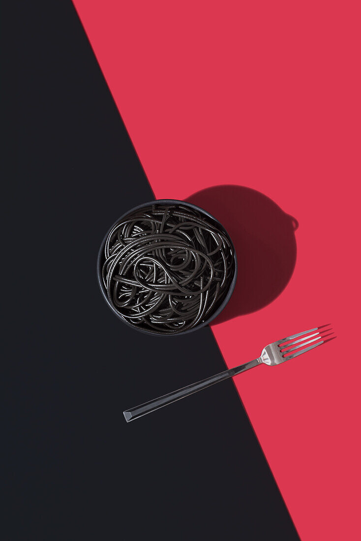 Draufsicht auf schwarze Spaghetti in einer Schüssel auf schwarzem und rotem Hintergrund neben einer Gabel