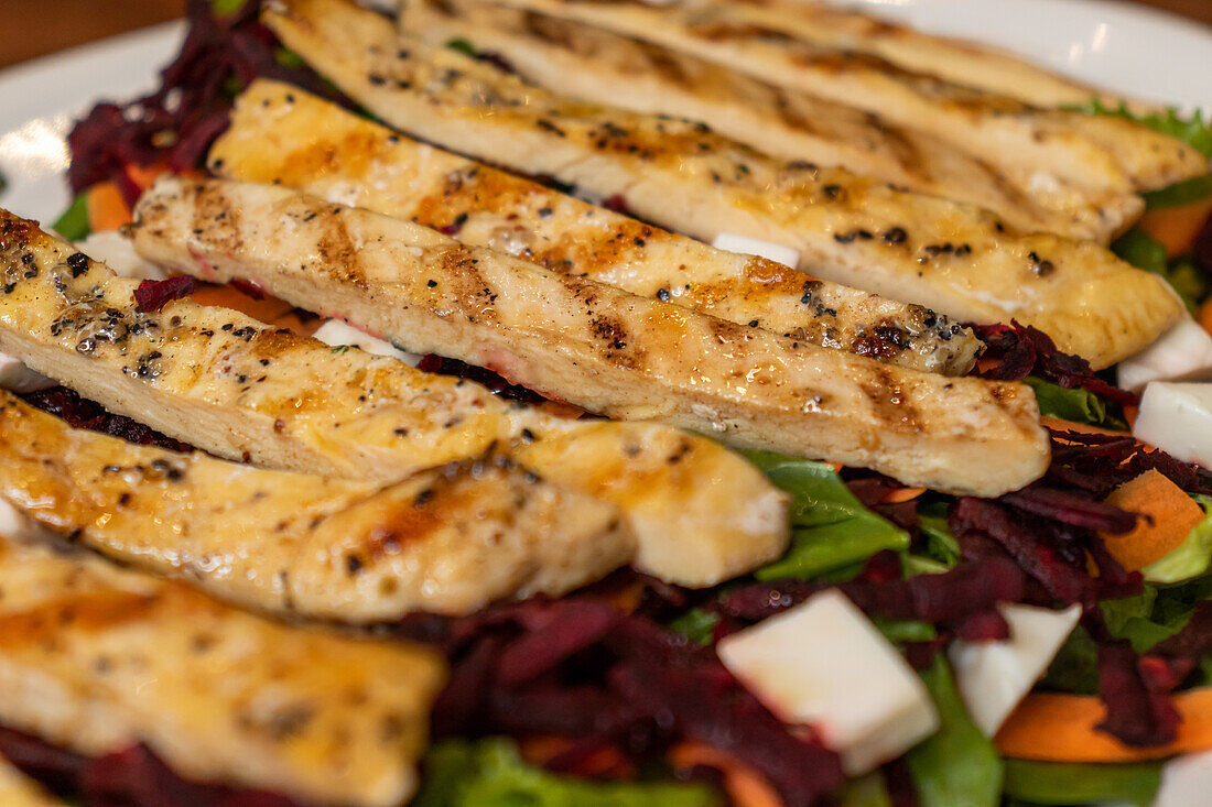 Nahaufnahme von gegrillter Hähnchenbrust auf grünem Salat mit Käsewürfeln, Karotten und gehacktem Rote-Bete-Gemüse, serviert auf einer weißen Marmorplatte im Licht