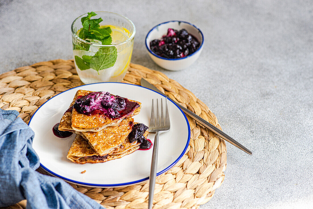 Stack of fresh homemade crepes topped with blueberry jam and a dollop of sour cream and lemon water drink, presented on a white plate with a wicker placemat.