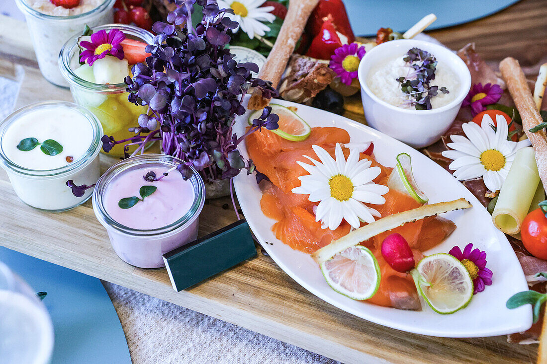 A colorful gourmet brunch arrangement featuring smoked salmon, edible flowers, and various dips served elegantly on a wooden board.