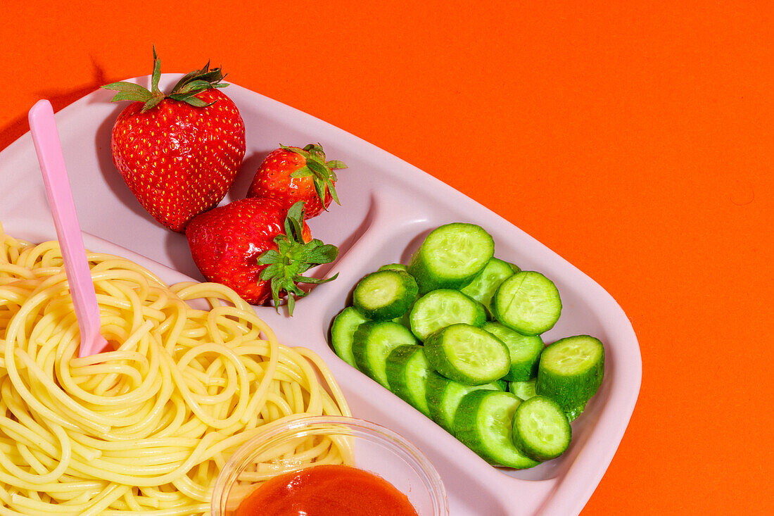 From above composition of spaghetti with tomato sauce cucumber slices and yummy fresh strawberries served for school lunch on orange top table