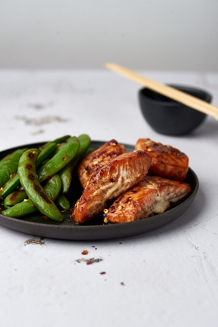 Juicy grilled chicken wings and snap peas on a black plate with chopsticks and a bowl in the background.