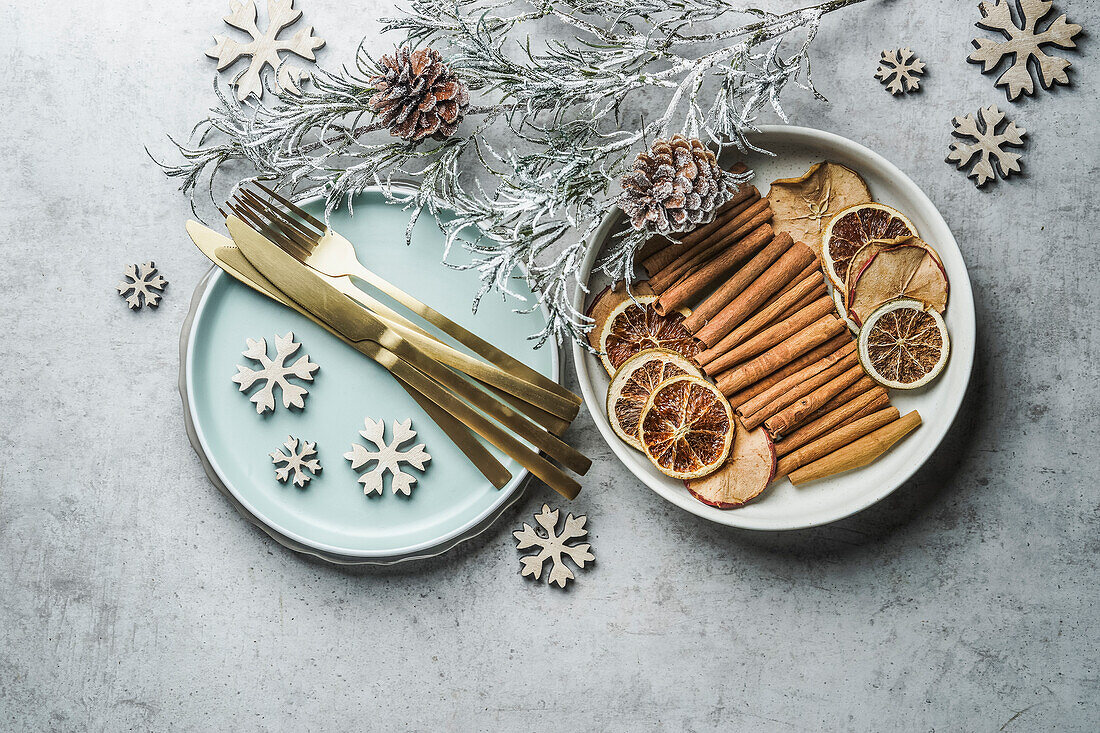 Weihnachtlicher gedeckter Tisch mit goldenem Besteck, hellblauem Teller, Zimtstangen, getrockneten Orangen und natürlichen getrockneten Äpfeln und Orangen und Winterzweigen auf grauem Tisch. Ansicht von oben