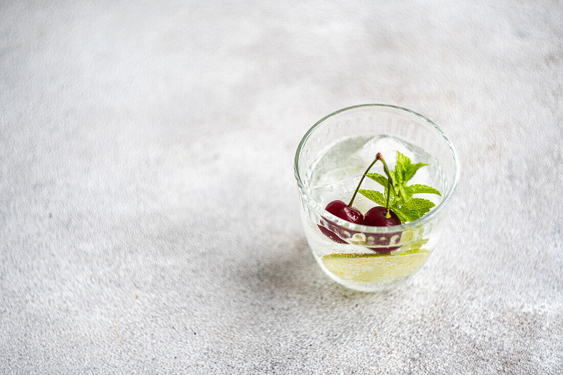 Draufsicht auf ein transparentes Glas mit einem erfrischenden Kaltgetränk mit Limette, Kirsche und Minzblättern auf einer grauen Fläche an einem Sommertag vor einem unscharfen Hintergrund