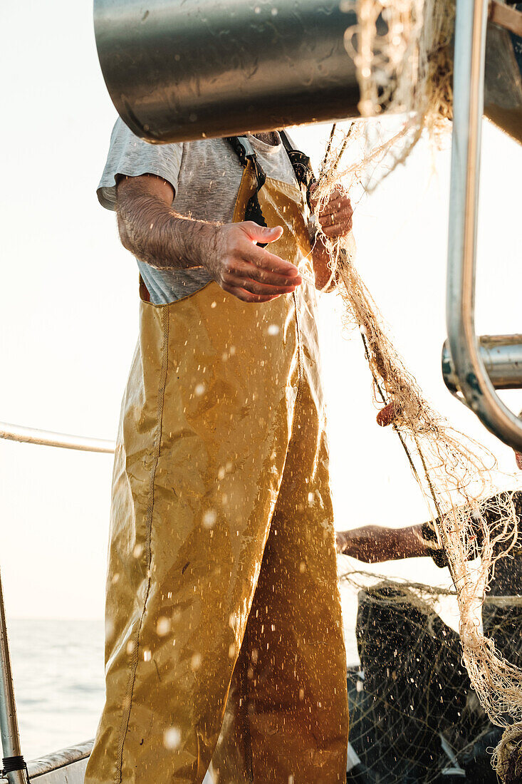 Seitenansicht eines abgeschnittenen, nicht erkennbaren männlichen Fischers in Uniform, der mit einem Netz auf Fischfang geht, während er auf einem Schoner in Soller nahe der Baleareninsel Mallorca arbeitet