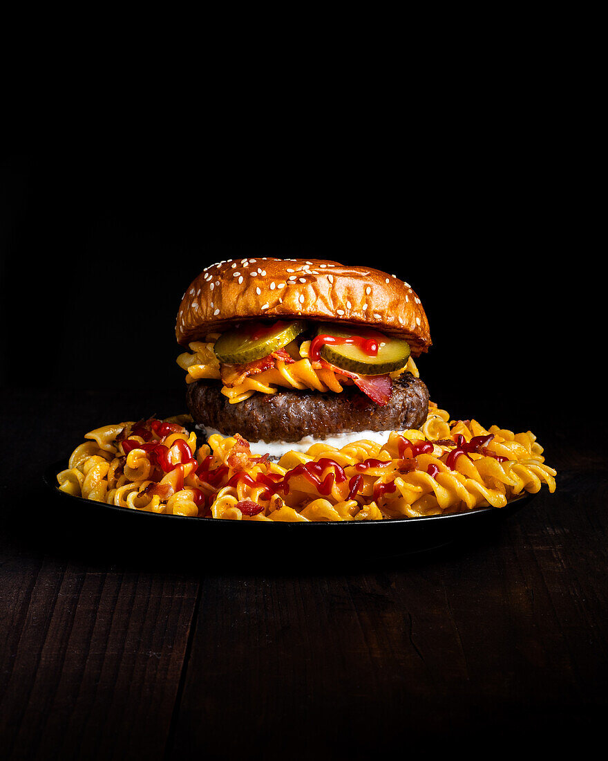 Hamburger auf einem Teller zwischen Makkaroni, Ketchup und Käse vor dunklem Hintergrund