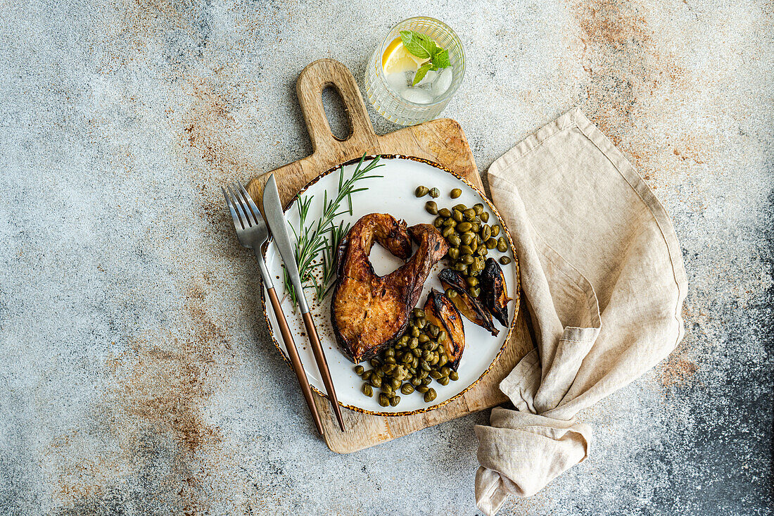 Top view of healthy grilled salmon steak seasoned with spices next to roasted lemons and capers on a wooden board with a linen napkin and lemon water