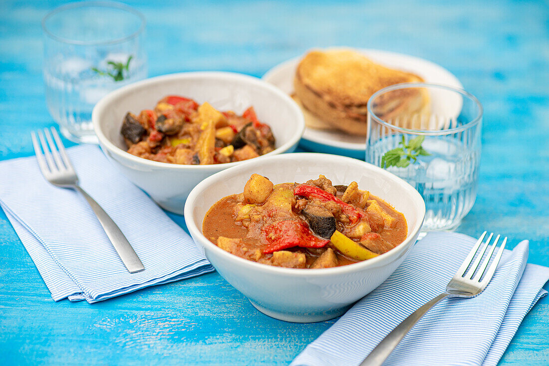 Traditional seasonal Georgian dish - Adjapsandali with stewed vegetables like potato, bell peppers, tomato, egg plant, coriander and Georgian spices placed on blue table near napkin and glass with water