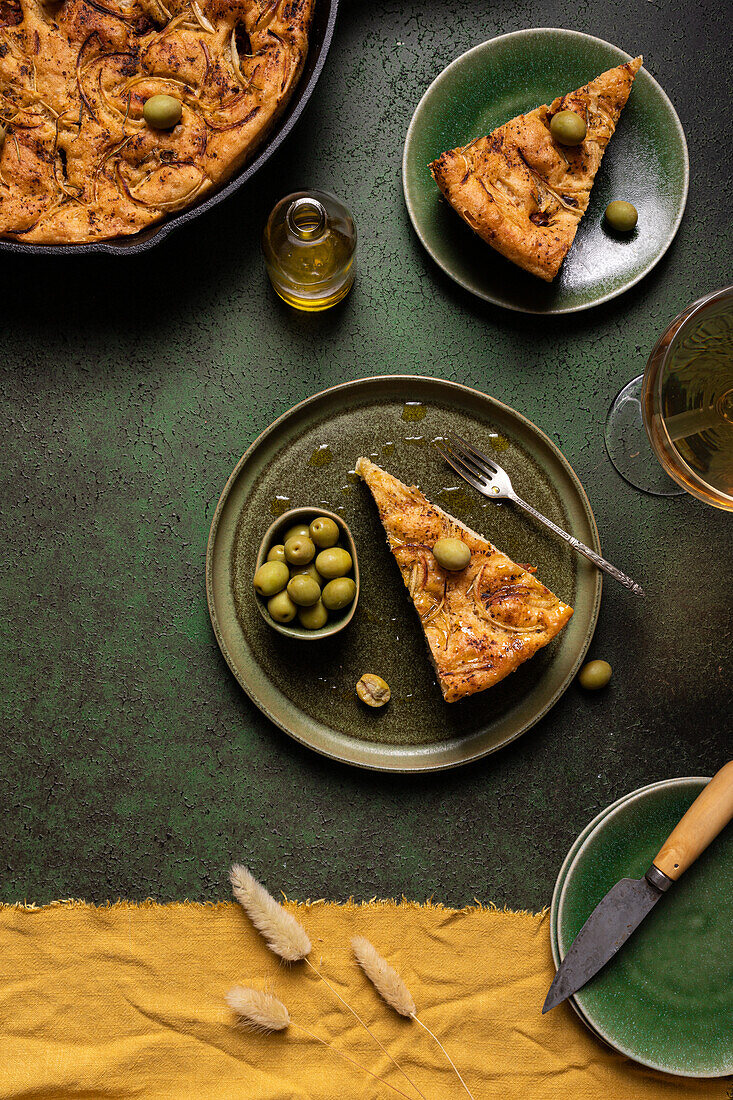 Draufsicht auf eine Scheibe gebackenes italienisches Focaccia-Brot mit grünen Oliven in einem Teller auf einem Esstisch mit Getränk auf grünem Hintergrund