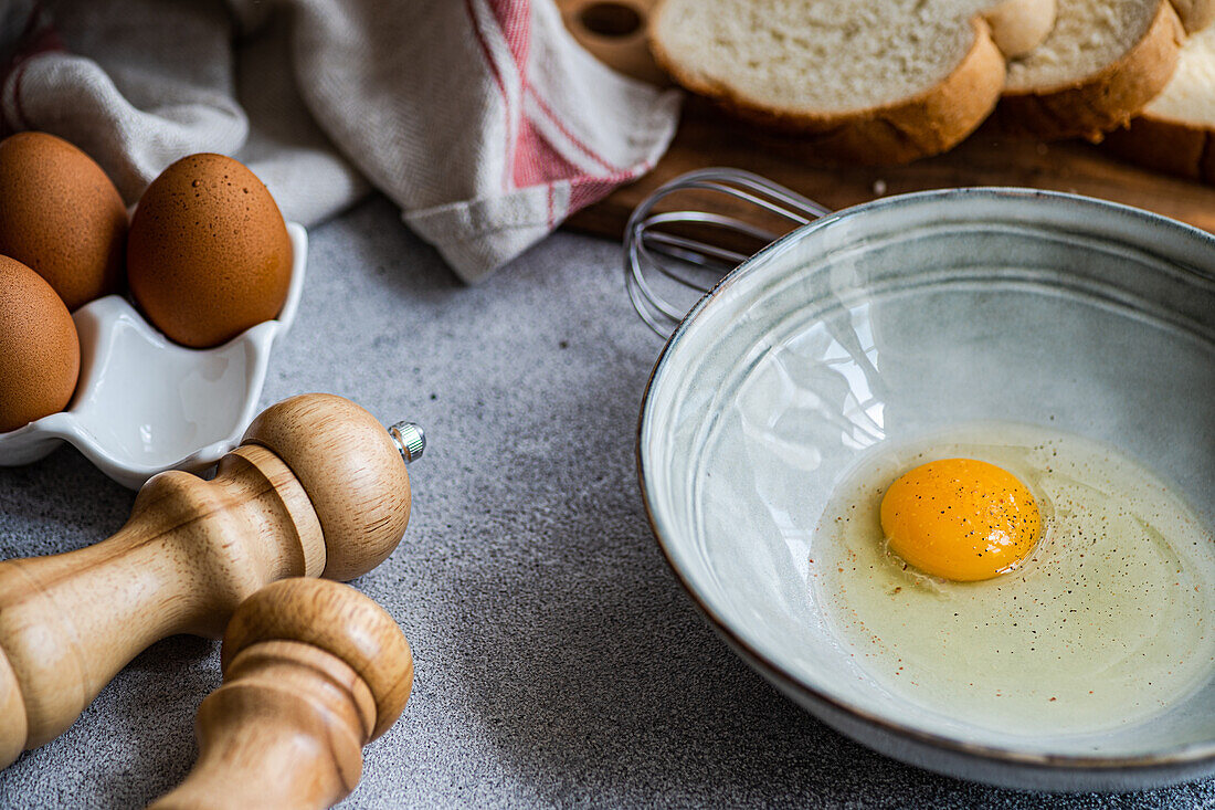 Raw egg in bowl near cutting board with fresh bread while cooking at concrete table with various ingredients in kitchen