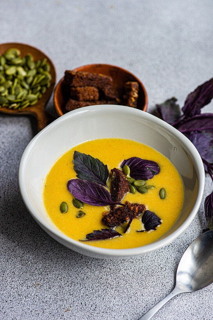 High angle of bowl with pumpkin cream soup with basil herb, rye bread and seeds on blurred grey background with leaves