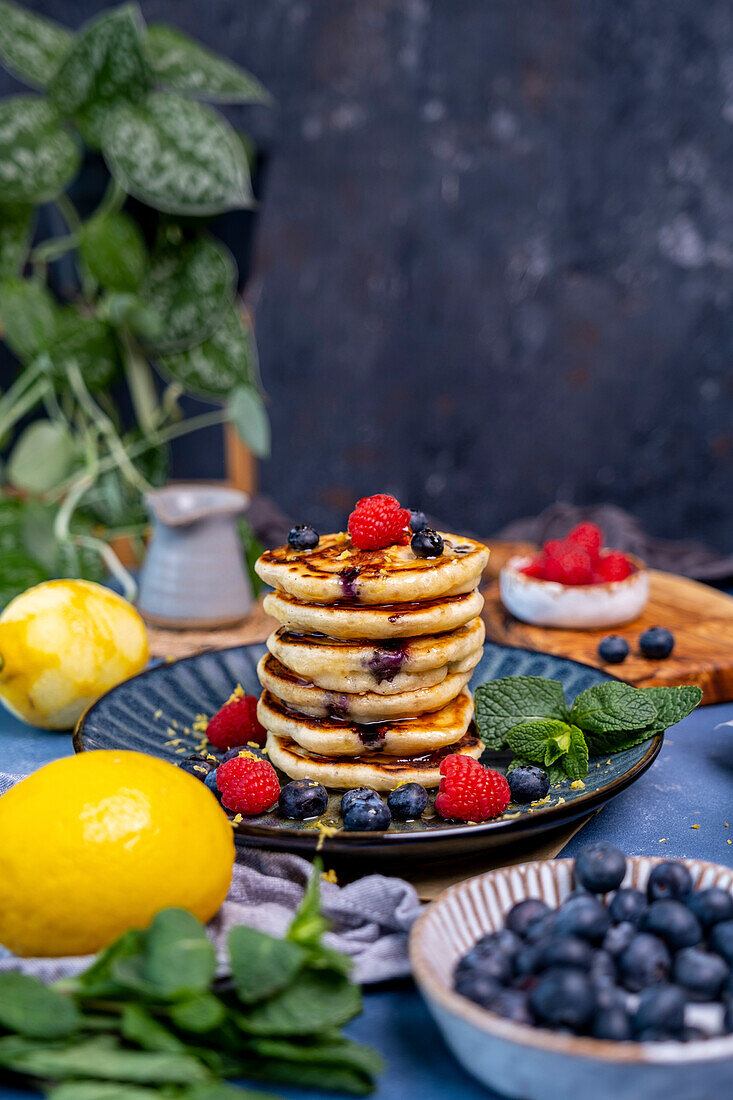 A stack of pancakes garnished with raspberries, blueberries and fresh mint on a dark background. Lemons, blueberries and raspberries around them