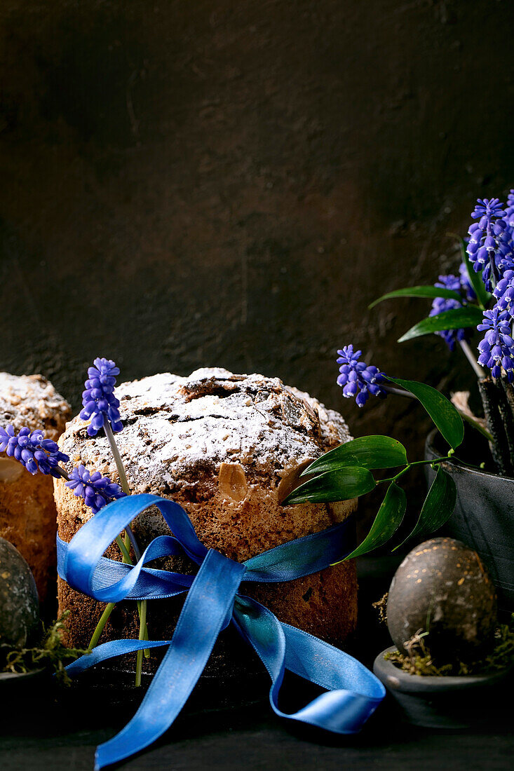 Hausgemachter traditioneller Oster-Panettone-Kuchen mit gefärbten schwarzen Eiern, blühenden Muscari-Blumen auf schwarzem Holztisch. Traditionelle italienische Osterbäckerei, Kopierraum, Nahaufnahme