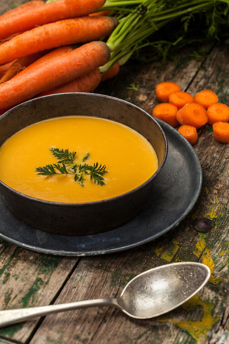 Eine Schüssel mit cremiger Karottensuppe, garniert mit frischen Kräutern, umgeben von ganzen und geschnittenen Karotten auf einem Holztisch