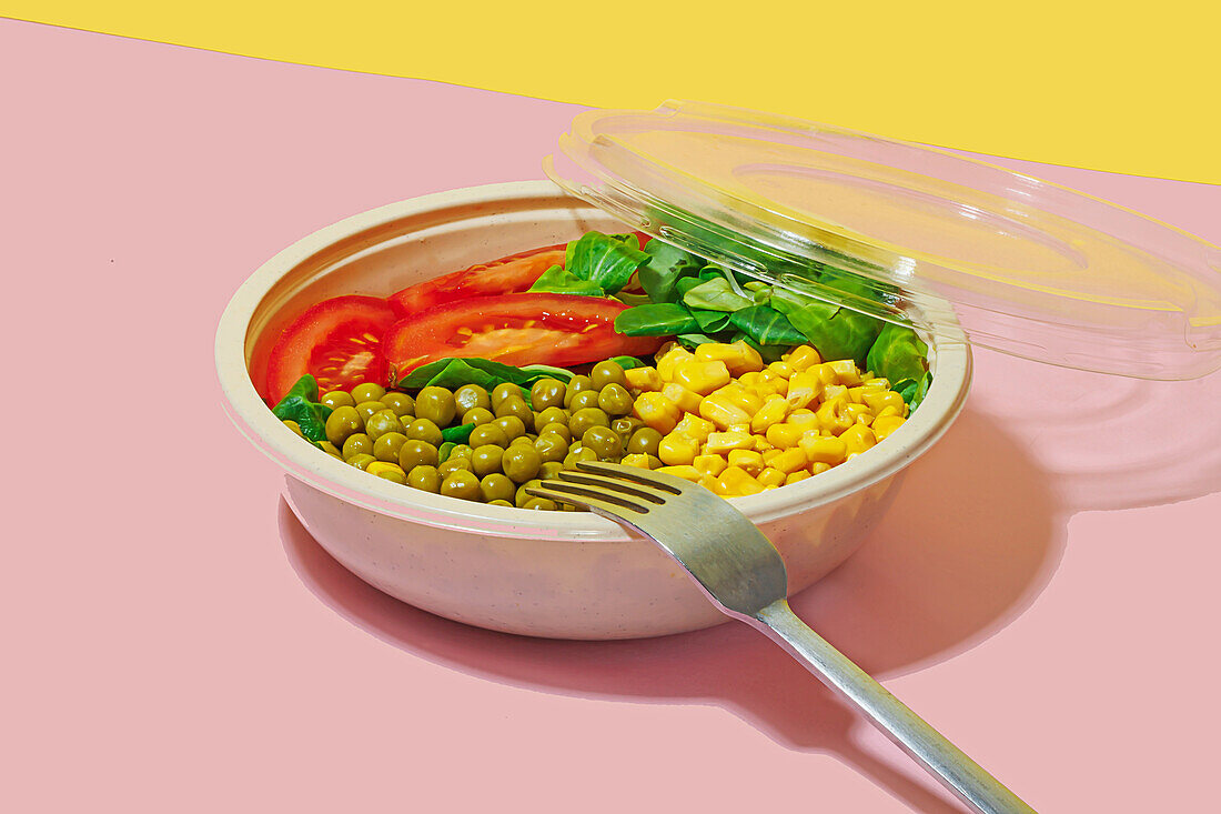 High angle of salad bowl with slices of tomato, spinach leaves, corn kernels and peas placed on pink surface with fork against yellow wall