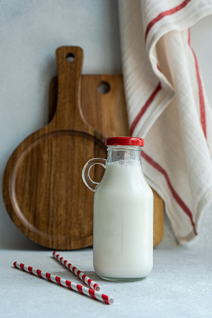 Rohe Kuhmilch in verschlossener Flasche neben Trinkhalmen auf grauer Fläche vor unscharfer Serviette und Schneidebrett