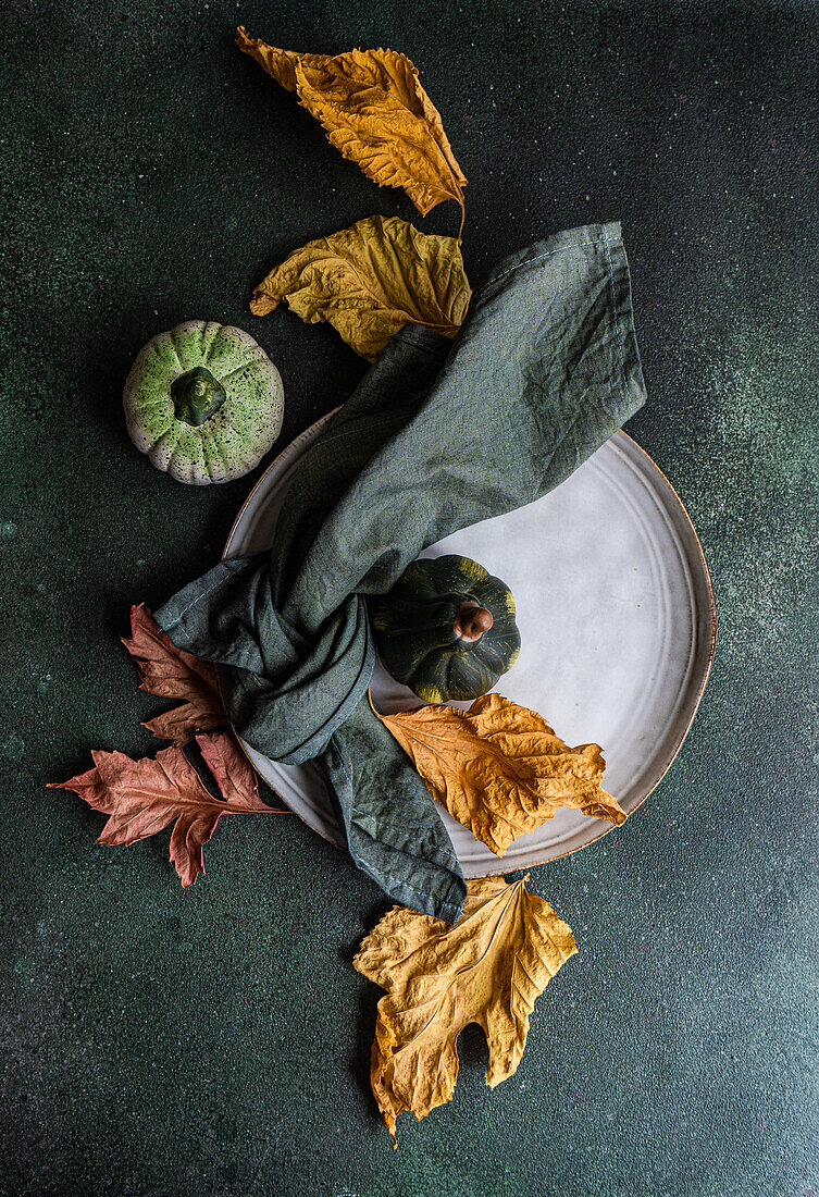 Top view of autumnal table setting with napkin, leaves and pumpkins placed on plate against dark surface