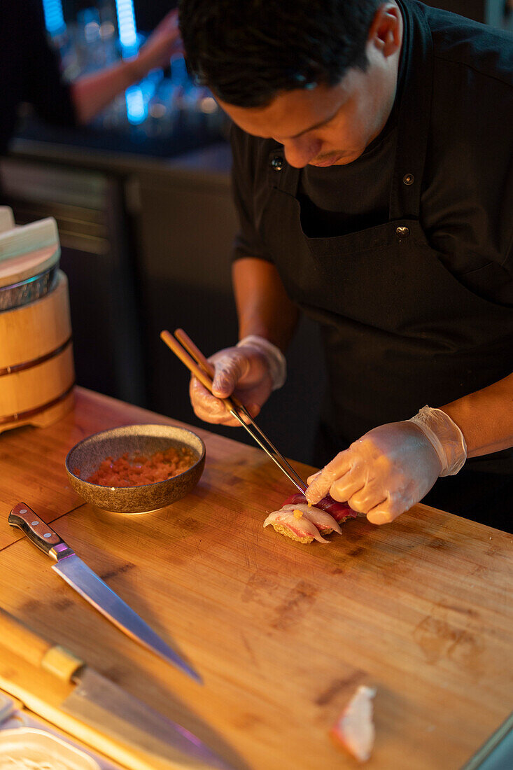 A sushi chef meticulously assembles a piece of nigiri sushi at a warmly lit restaurant counter, showcasing culinary precision.
