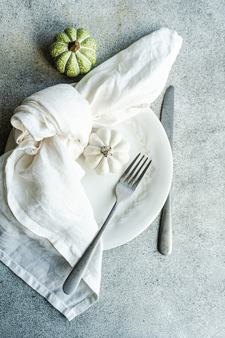 Top view of white ceramic plate with decorative pumpkins with silver fork and knife, wrapped in a white napkin, placed on a textured gray concrete surface