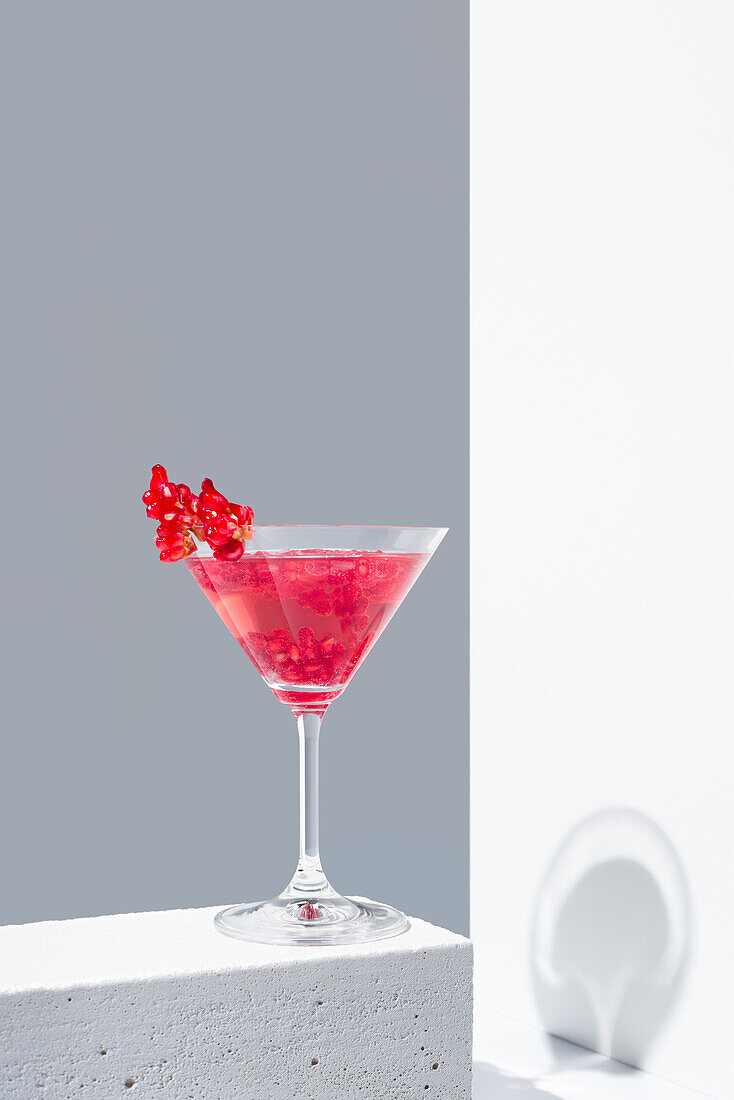 Glas mit rotem Granatapfelcocktail und Granatapfelkernen vor grauem und weißem Hintergrund, der Schatten wirft