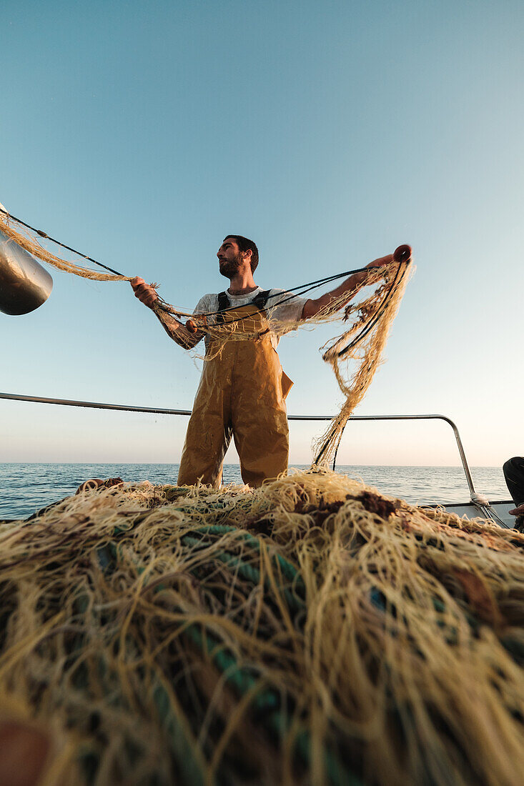 From below focused bearded male fisher in uniform seiner hunting fish with net while working on schooner in Soller near Balearic Island of Mallorca
