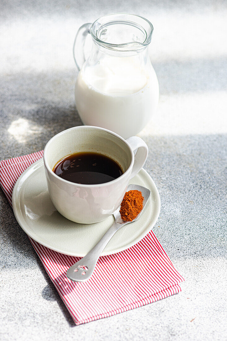 Hochformatige Tasse würziger Espresso neben Löffel mit roter Paprika auf grauem Tisch neben Milchflasche im Tageslicht