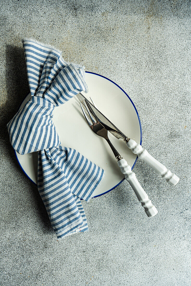 Draufsicht auf einen minimalistisch-rustikal gedeckten Tisch mit weißem Teller, Besteck und gestreifter Serviette auf grauem Untergrund