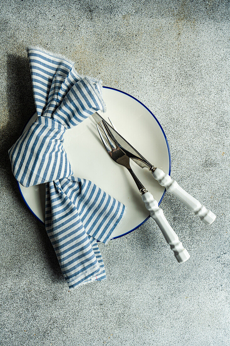 Top view of minimalistic rustic table setting with white plate cutlery and striped napkin on gray surface