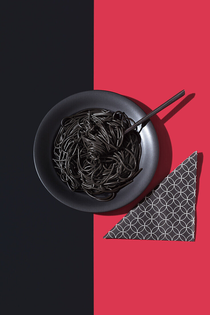 Draufsicht auf schwarze Spaghetti in einer Schüssel mit Gabel auf schwarzem und rotem Hintergrund neben einer Serviette