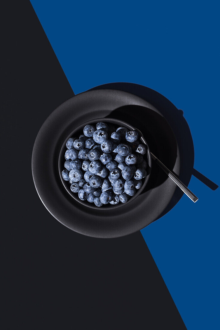 From above of bowl full of ripe blueberries accompanied by a spoon on a dual-tone blue and black background