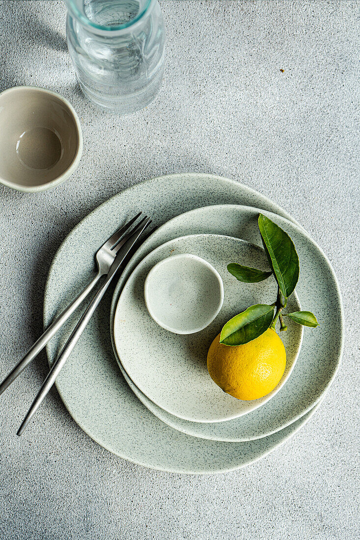 A vibrant lemon with leaves placed on a set of speckled ceramic plates, accompanied by sleek cutlery and glassware on a textured surface.