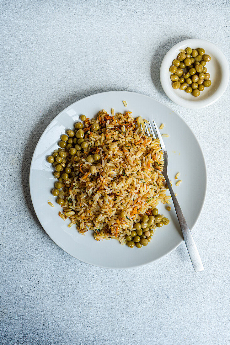 Draufsicht auf gekochten Reis mit Zwiebel, Karotte, Paprika, grünen Erbsen und Gewürzen auf Teller mit Gabel neben Teller mit grünen Erbsen vor grauem Hintergrund