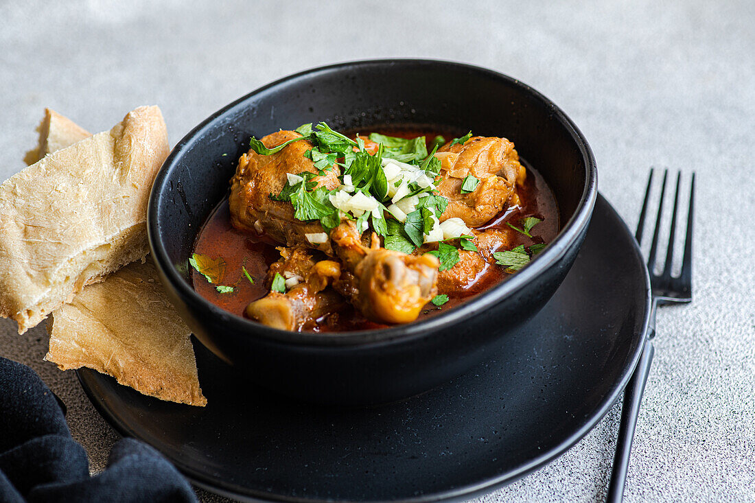 High angle of Georgian dish Chakhokhbili is stewed chicken meat in tomato sauce with spices and herbs served on black plate with some bread on gray table near fork and napkin against blurred background